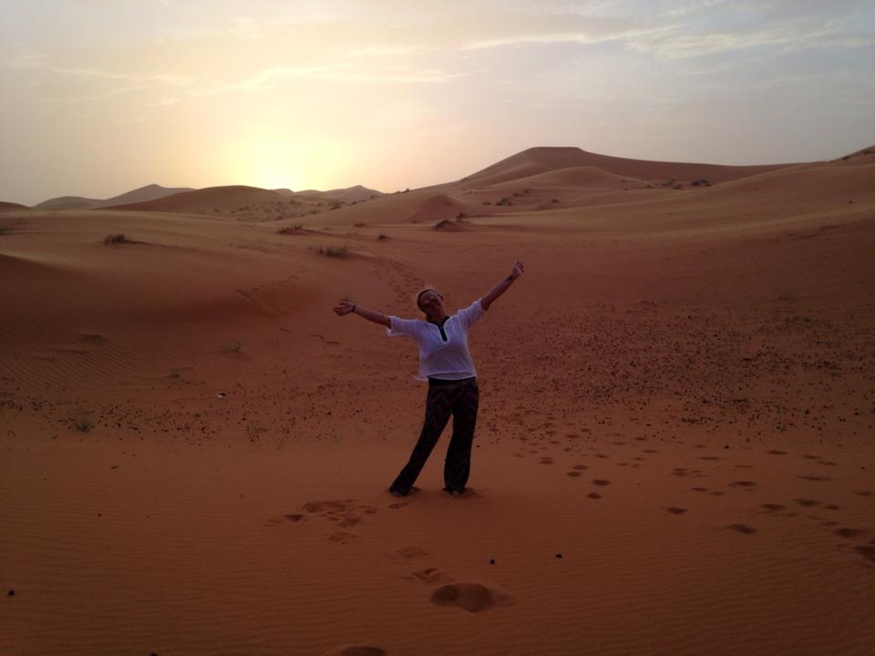Me, a young woman traveler, posing in a wide landscape of nothing but sand in the Sahara Desert in Morocco at sunset.